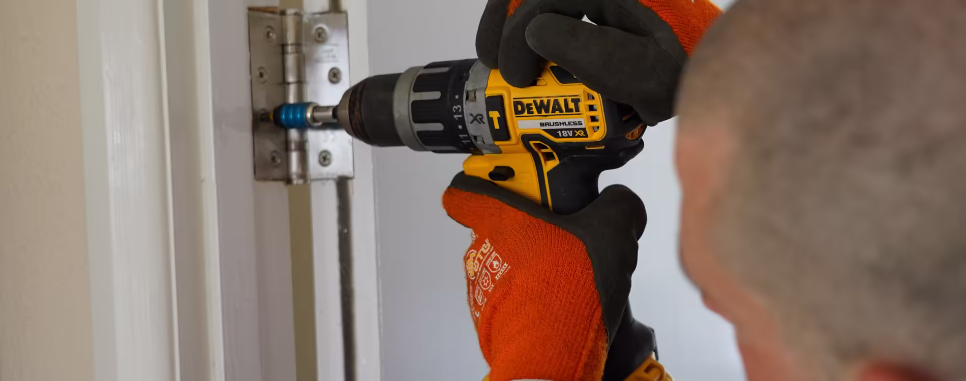 A close-up of a technician's hands wearing orange and black gloves, using a yellow DEWALT cordless drill to tighten or remove a screw on a metal door hinge, illustrating a responsive repairs service for a residential property.