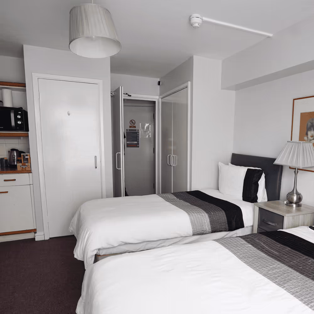 A newly refurbished resident's twin room in a care facility, featuring fresh white decor, two beds with modern bedding, and a compact kitchenette area.