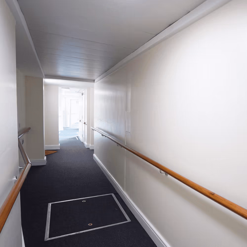 A long communal corridor after redecoration, with clean white walls, new dark carpeting, and a continuous wooden handrail for resident support.
