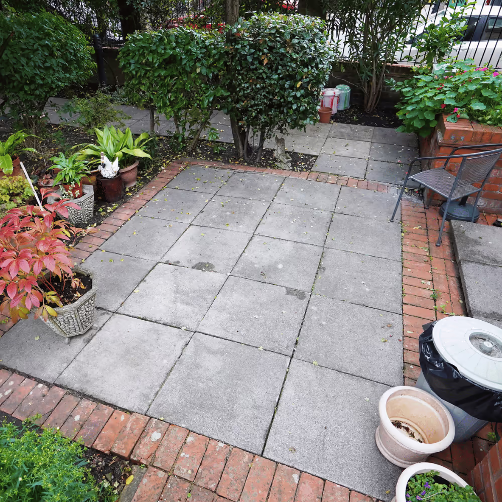 A neatly laid patio area in the communal garden, featuring grey paving slabs with a red brick border, surrounded by residents' potted plants.