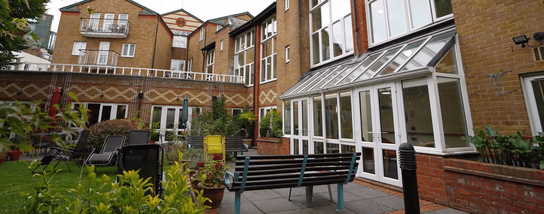 The exterior courtyard of a residential care facility, showing a new conservatory and a pleasant patio garden with seating for residents after a communal repairs project.