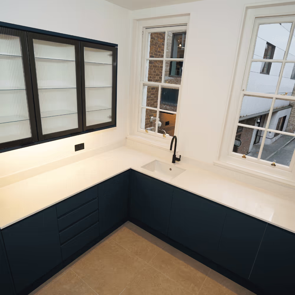 A high-end, modern office kitchen created during a refurbishment, with stylish dark blue cabinets, white countertops, reeded glass wall units, and traditional sash windows.