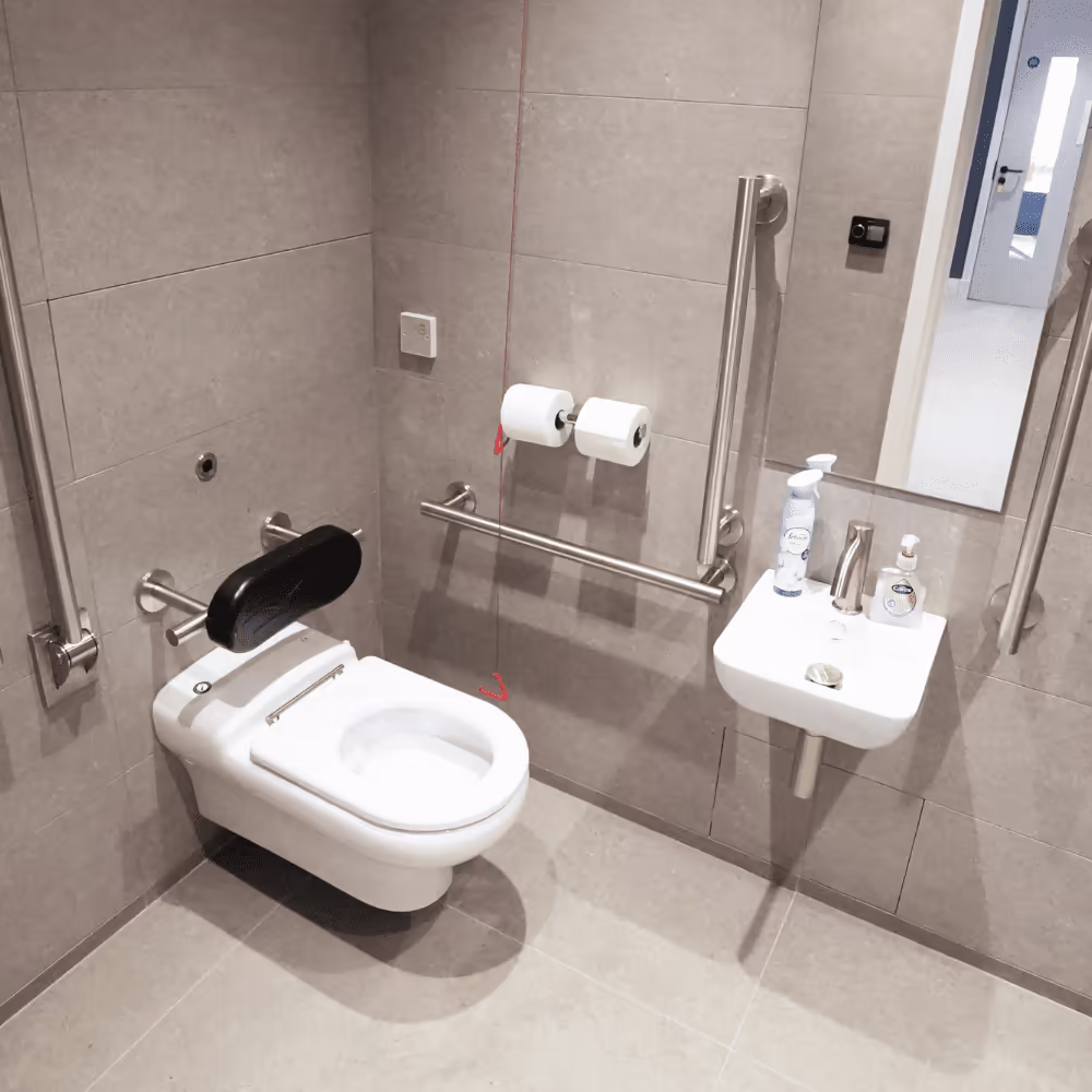 A newly installed, fully accessible WC in a refurbished office, featuring modern grey tiles, a wall-hung toilet with a backrest, and compliant stainless steel grab rails.